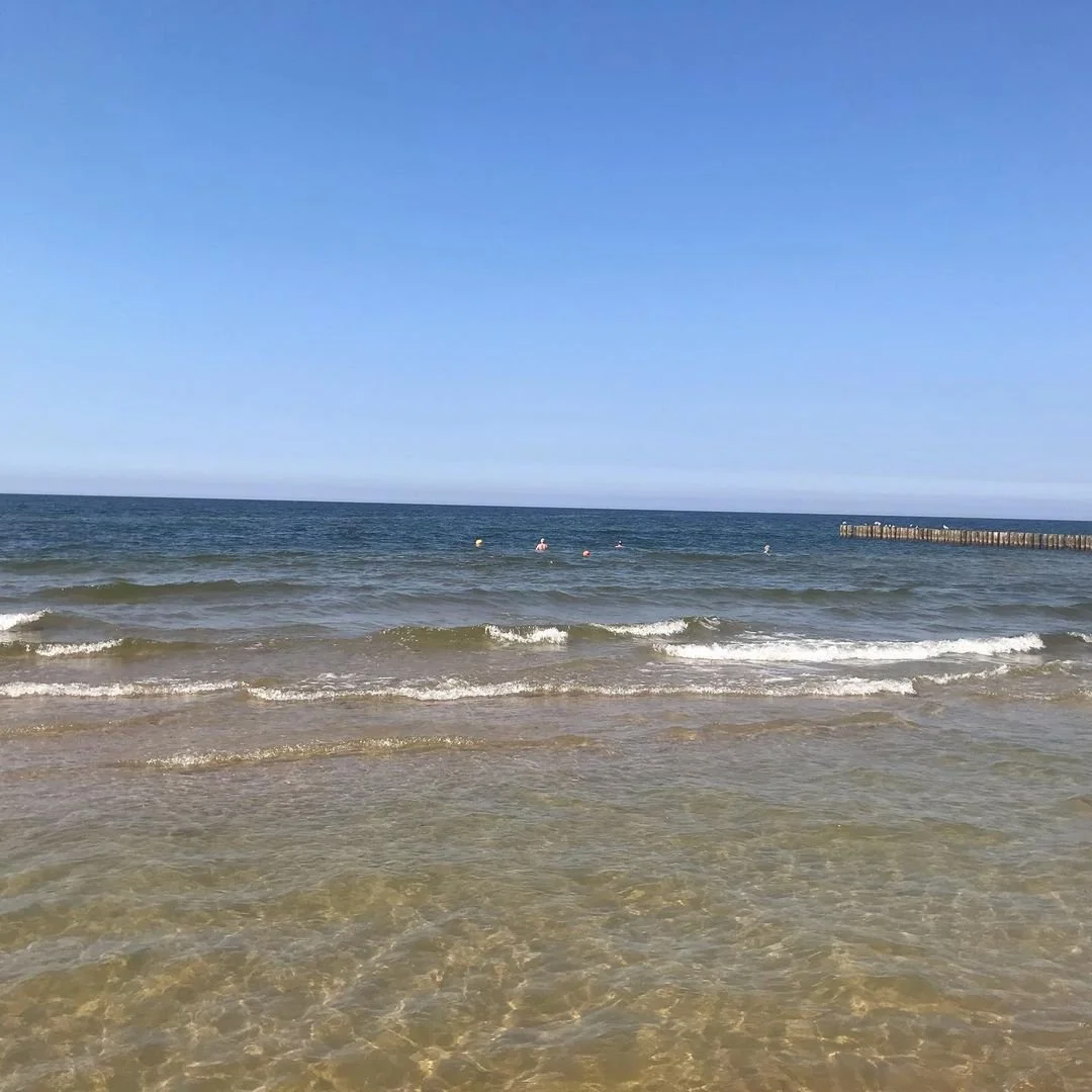 Sunny beach with small waves, ocean water, and a clear blue sky, with a few people swimming and a pier on the right side.