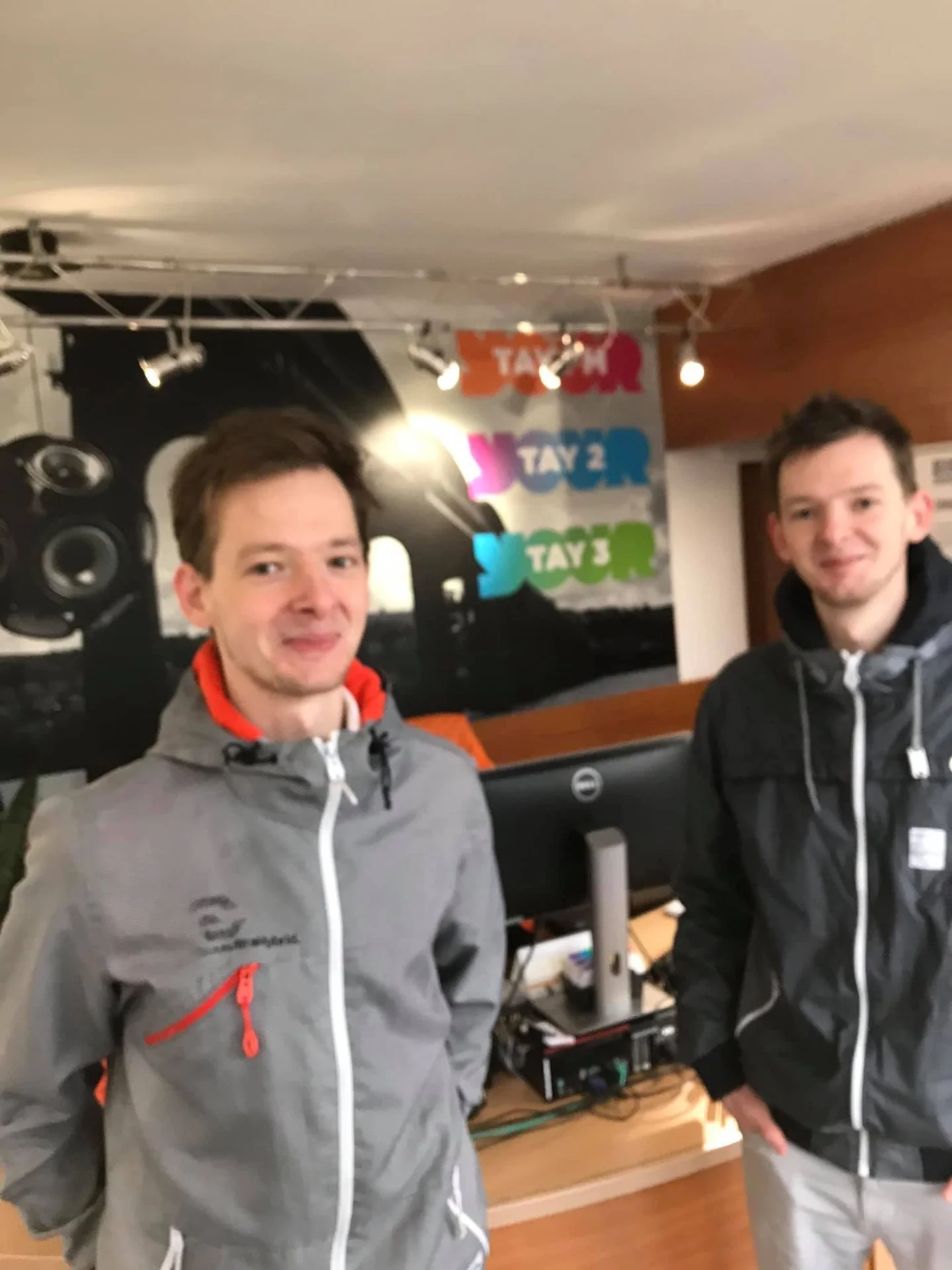 Two young men standing indoors in front of a colorful wall with text, wearing outdoor jackets.