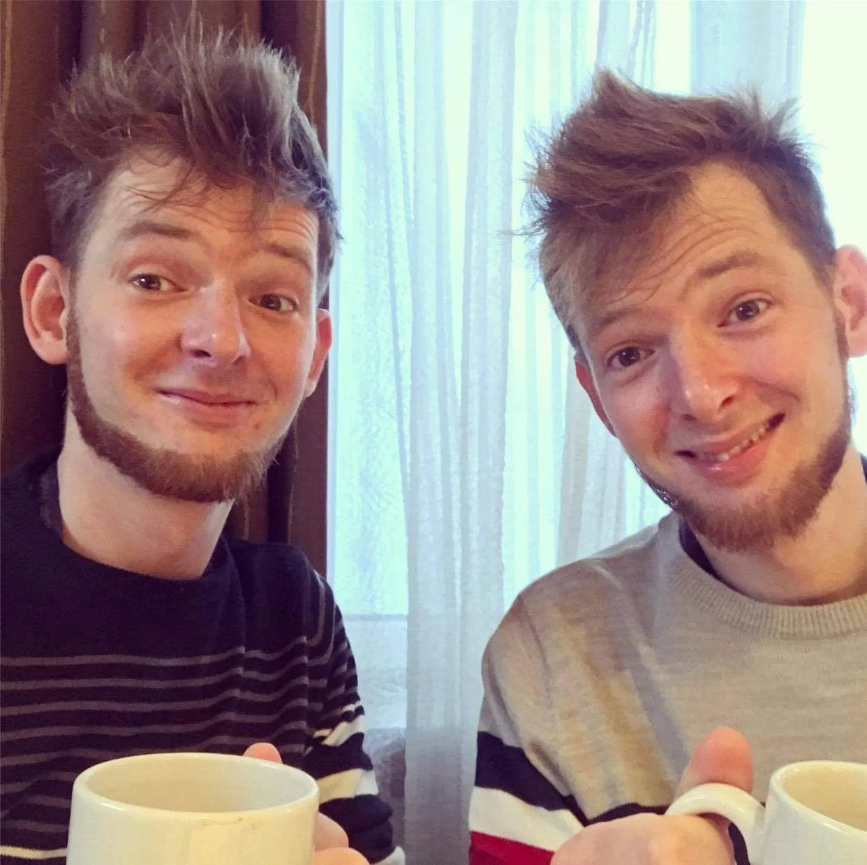 Two young men with beards and styled hair smiling while sitting at a table with coffee mugs in front of them, in a room with a window with sheer curtains in the background.