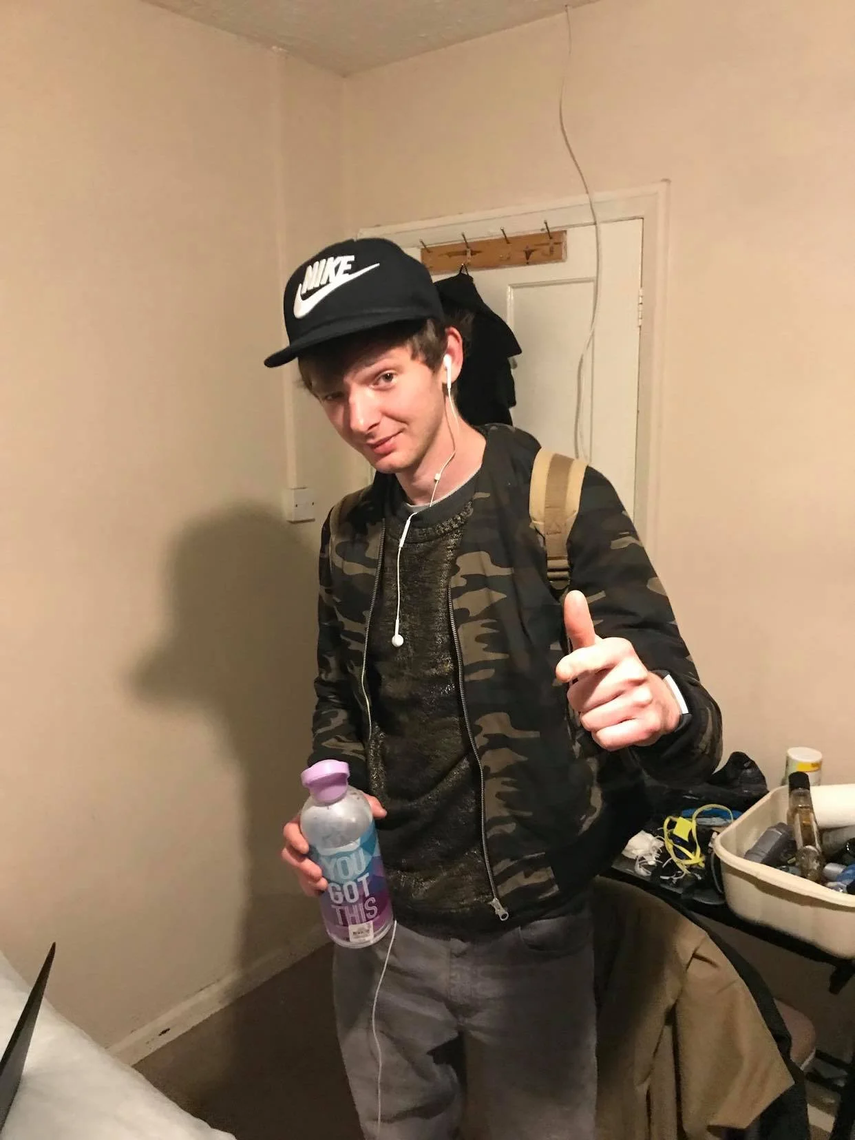 Young man with a Nike cap, camo jacket, carrying a backpack, holding a pink water bottle, giving a thumbs-up, and listening to earphones in a room with a table and various items.