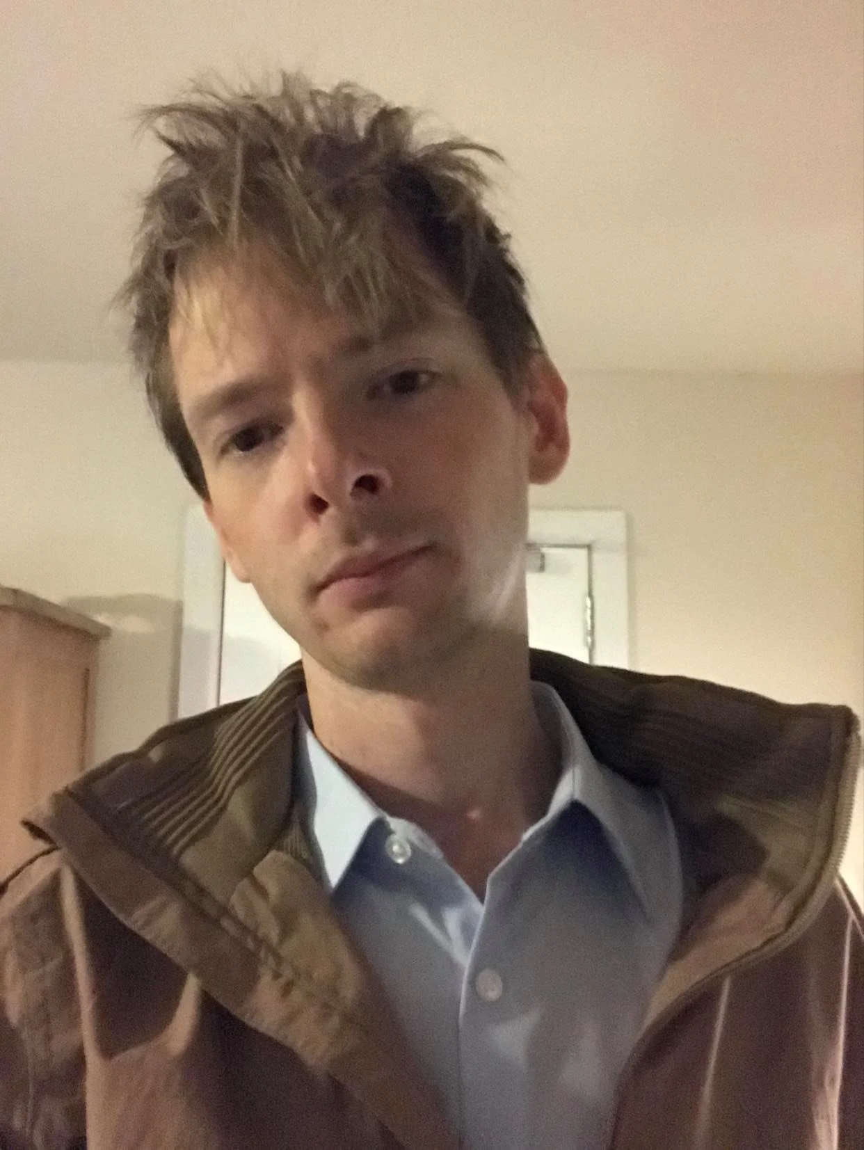 A young man with messy, light brown hair wearing a light blue collared shirt and a brown jacket, indoors with a plain background.