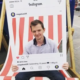 A young man sitting in front of a red and white striped background, holding a large Instagram photo frame prop, smiling at the camera.