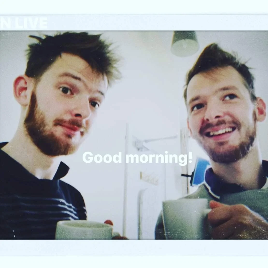 Two young men with beards smiling and holding coffee mugs in a bright room with a white wall. The text 'Good morning!' appears in the center.