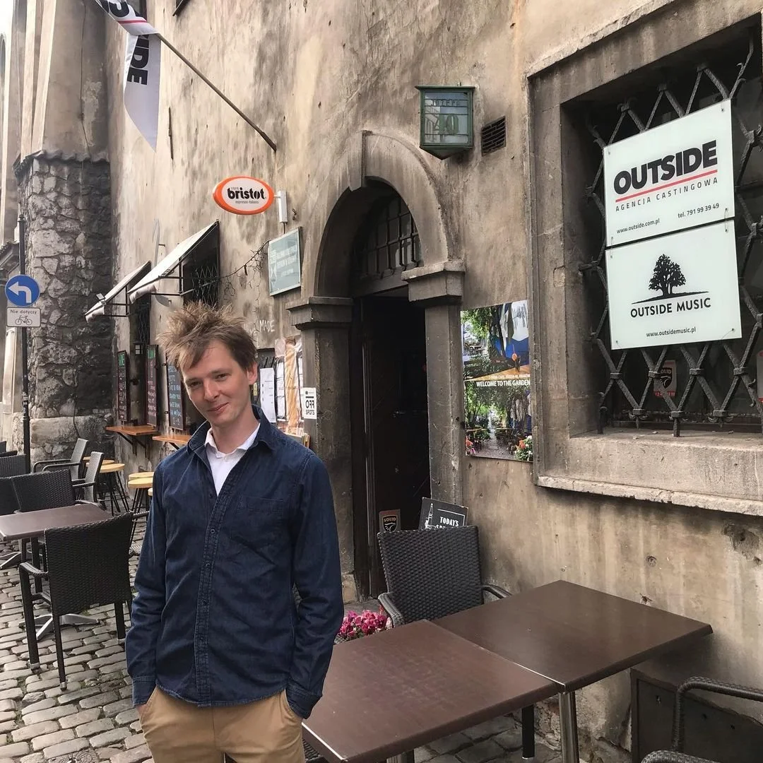A young man with light brown hair, wearing a dark blue jacket over a white shirt, standing outside a rustic cafe or restaurant with outdoor seating on a cobblestone street.