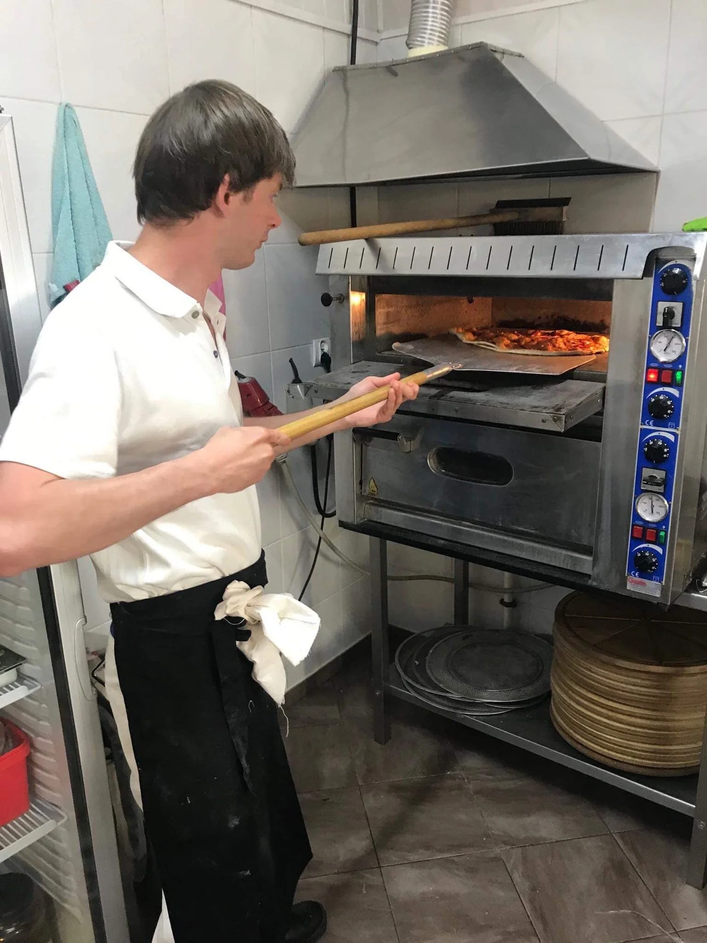 A man is using a pizza peel to place a pizza into a commercial pizza oven in a restaurant kitchen.