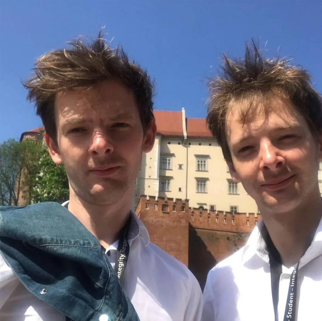 Two young men with spiky hair taking a selfie outdoors on a sunny day, with a building and trees in the background.