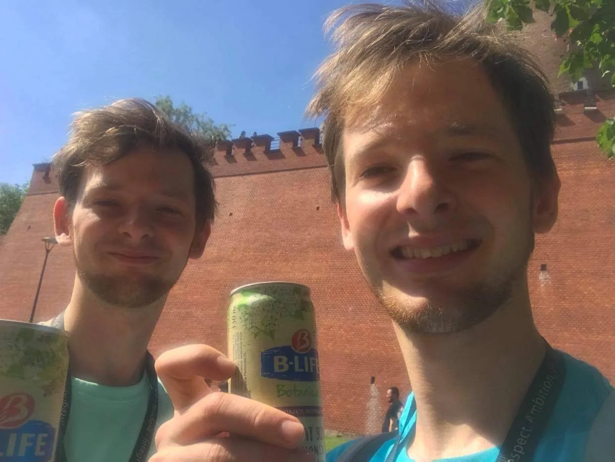 Two men smiling outdoors in front of a red brick wall, holding cans of B-LIFE beverage.