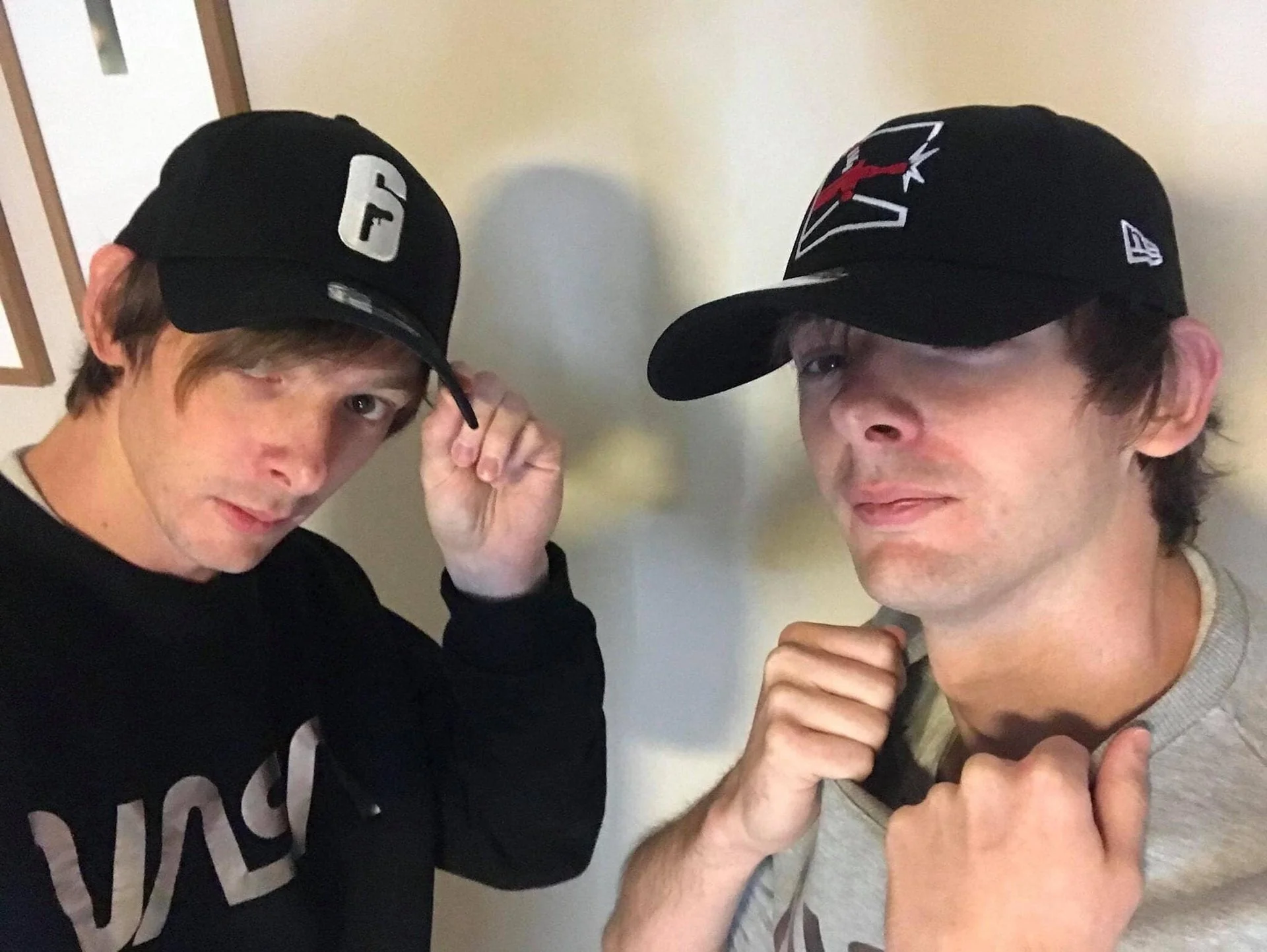 Two men wearing baseball caps posing indoors, with one adjusting his cap and the other holding his shirt collar.