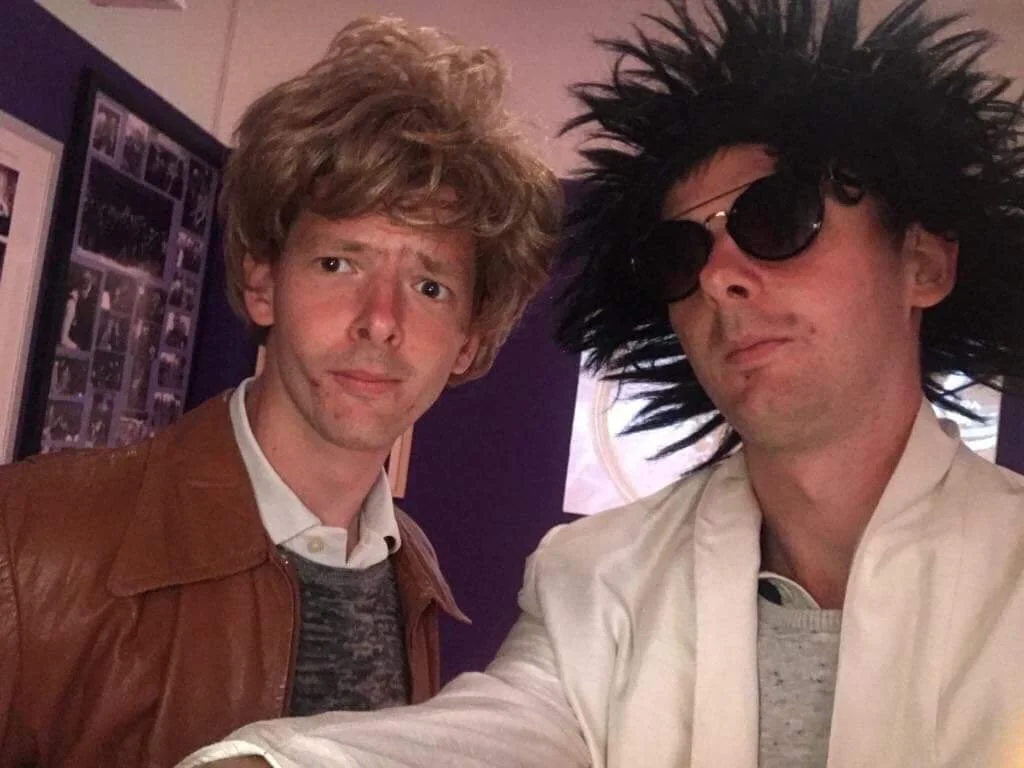Two men dressed in costumes, one with messy hair, leather jacket, and a confused expression, and the other with spiky wig, sunglasses, and a light-colored jacket.