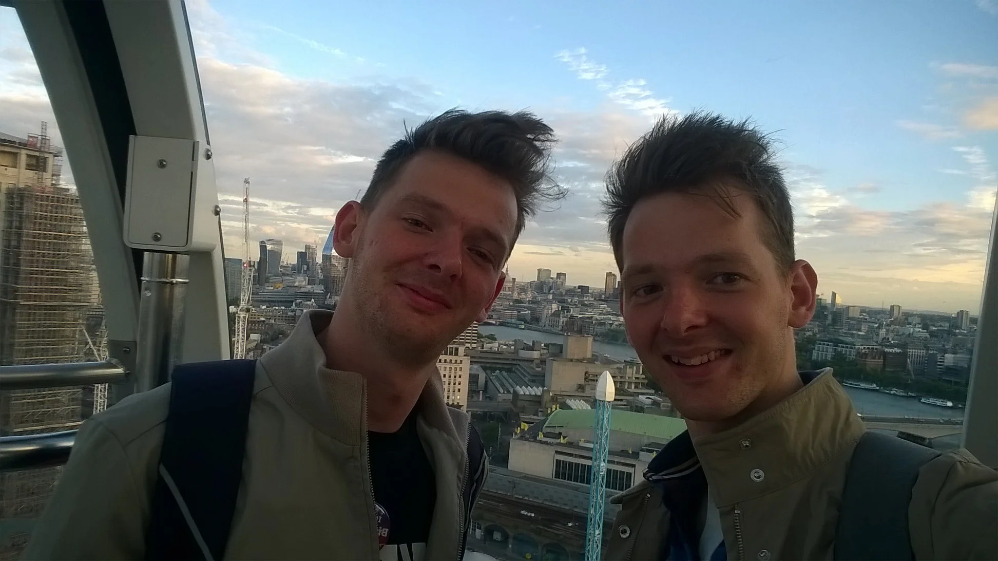 Two young men taking a selfie inside a Ferris wheel cabin with a city skyline, river, and partly cloudy sky in the background.