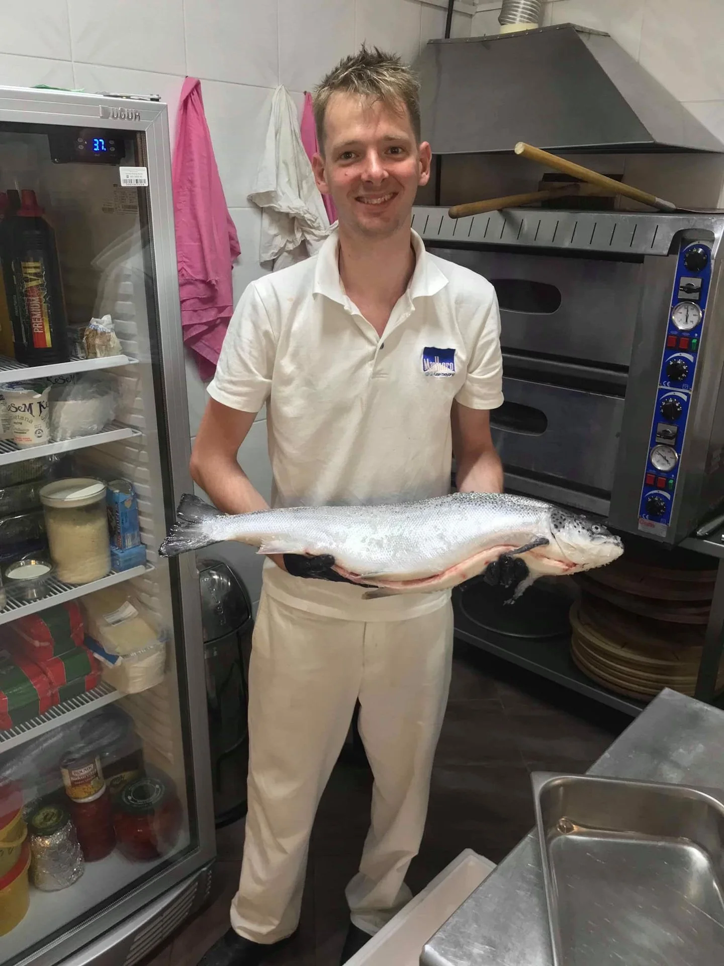 A man in a white shirt and pants holding a large fish in a kitchen.