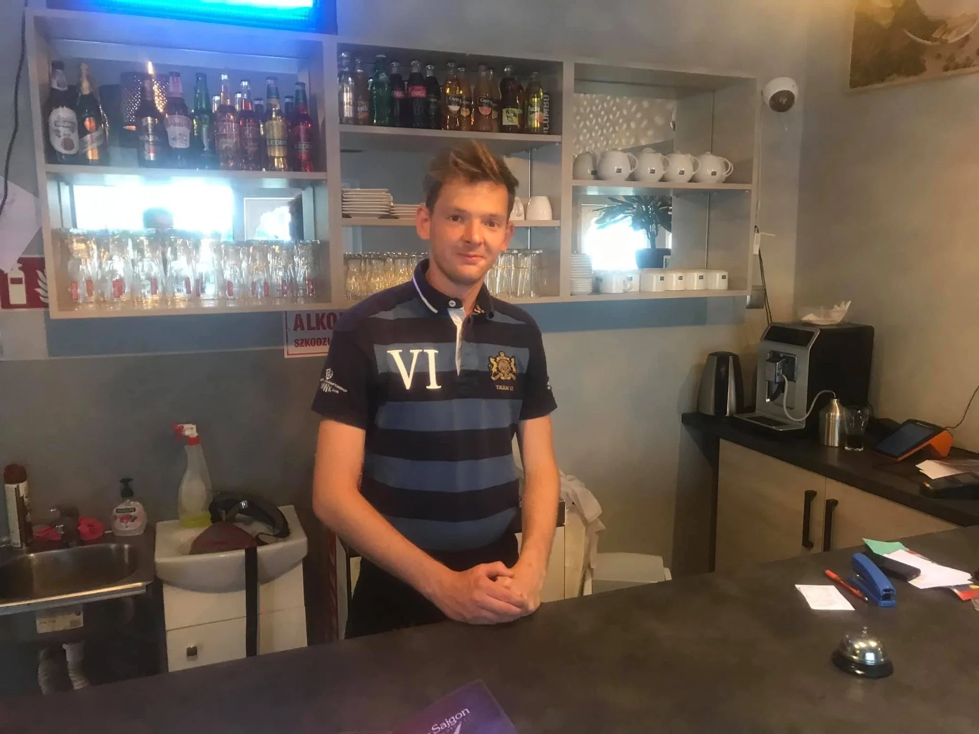 A young man with brown hair smiling, standing behind a bar counter in a cafe or bar setting, with shelves of bottles, glasses, and cups behind him, and various bar equipment on the counter and nearby
