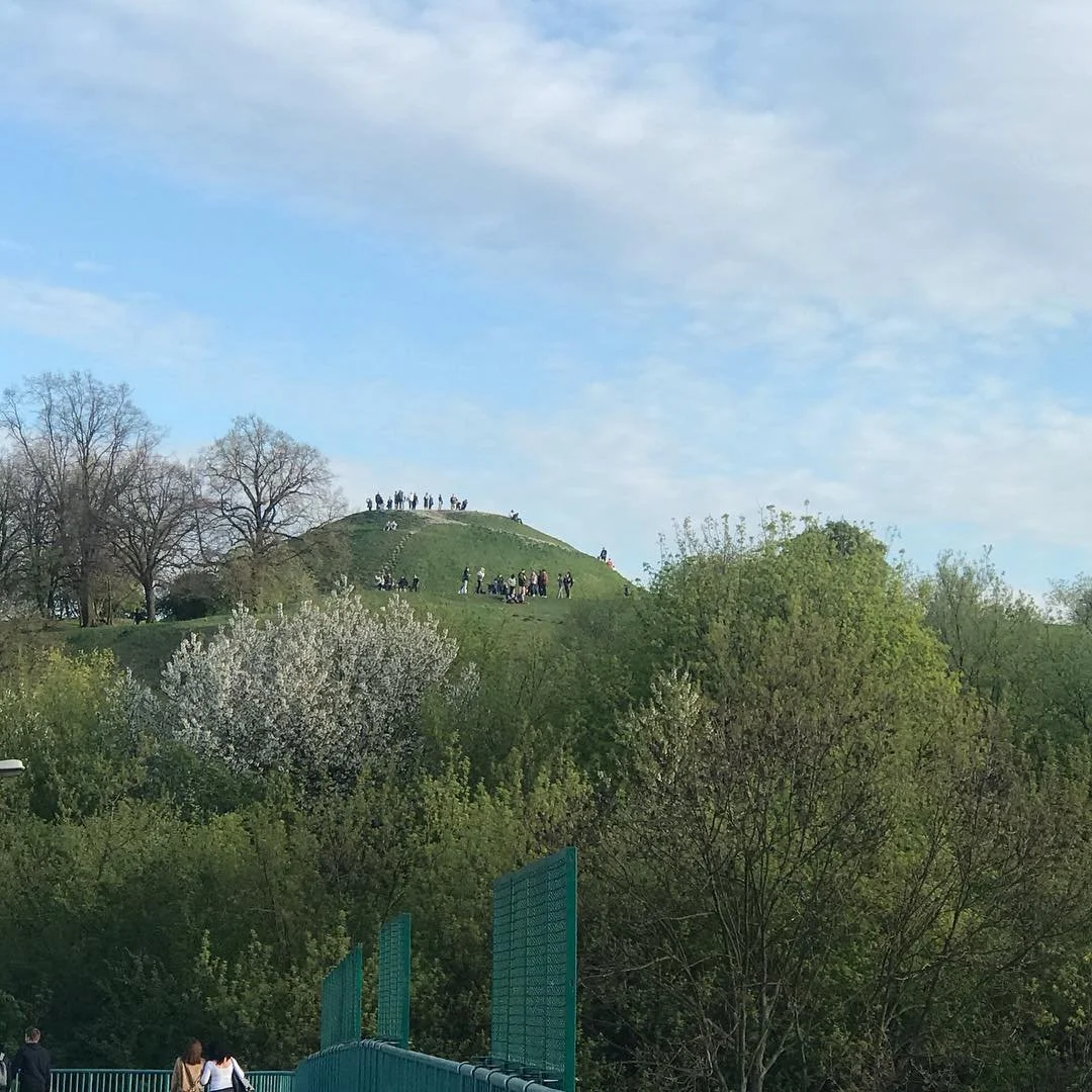 A grassy hill with people walking and standing on top and around it, surrounded by trees under a partly cloudy sky.