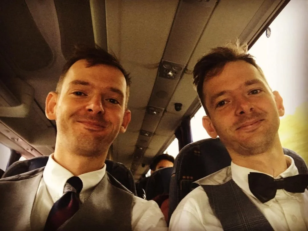 Two men in formal attire sitting on a bus, smiling at the camera. They are wearing dress shirts, vests, and one with a bow tie. The background shows the bus interior with other passengers.