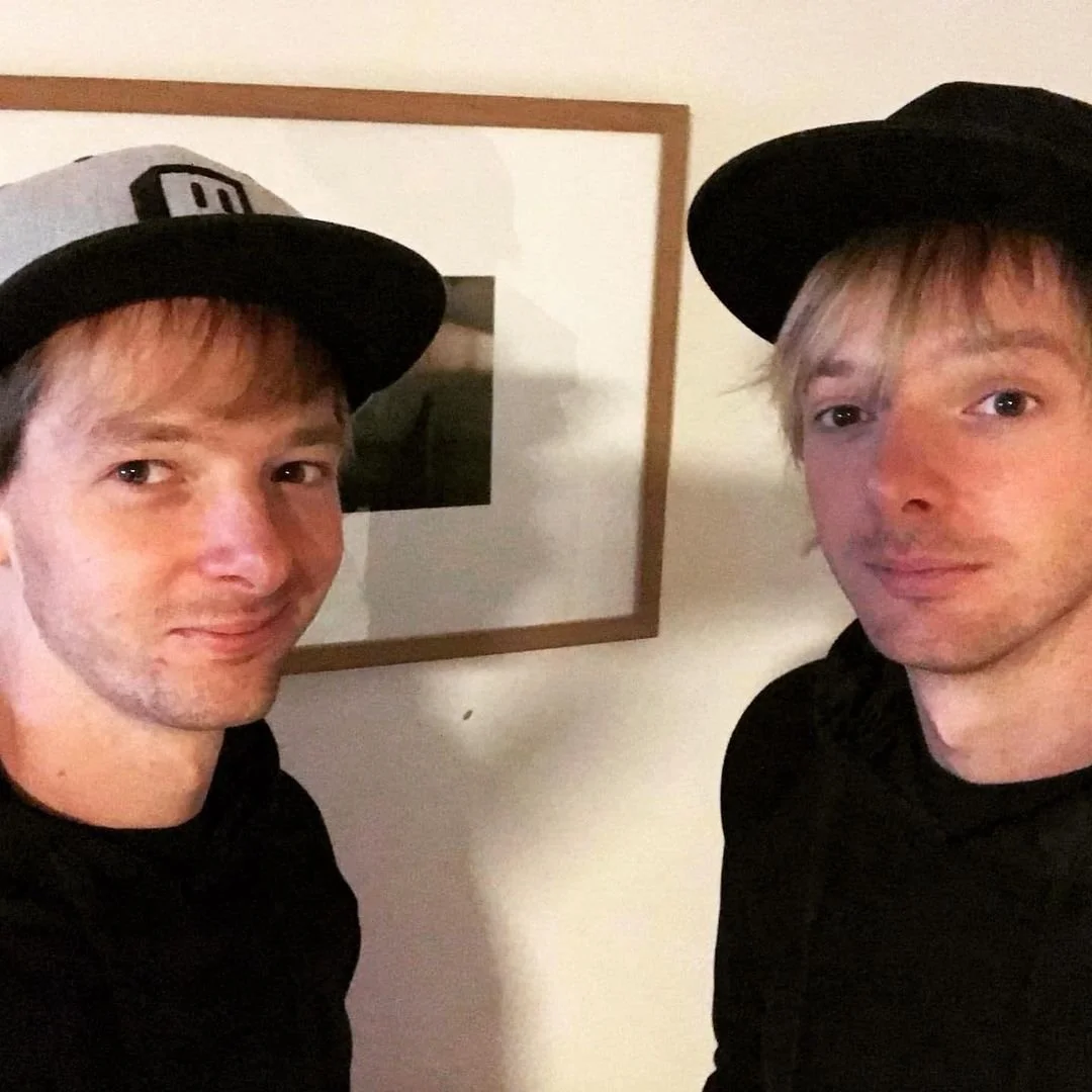 Two young men wearing black hoodies and baseball caps, standing indoors in front of a white wall with a framed picture.
