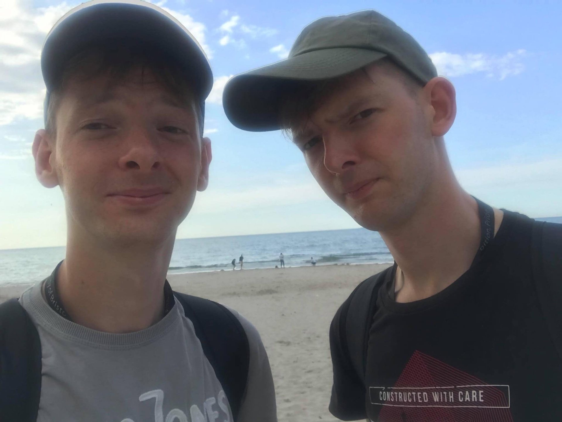 Two young men wearing caps take a selfie on a beach with the ocean and a cloudy sky in the background. There are some people walking on the beach in the distance.