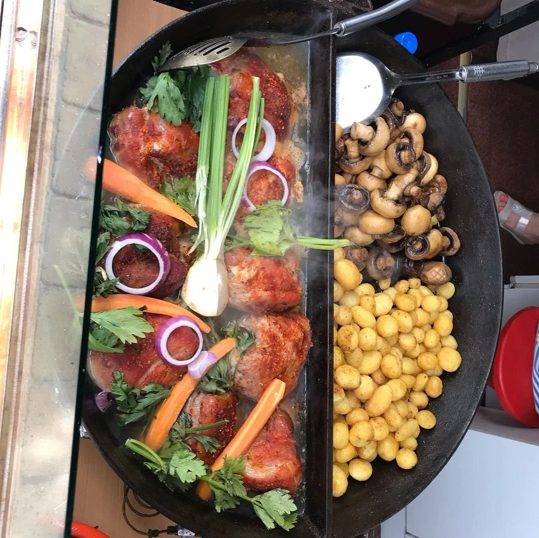 A large black skillet with seasoned chicken leg quarters topped with herbs, onions, and carrots, on the left side. The right side of the skillet holds sautéed mushrooms and tiny potatoes. Several cooking utensils are placed on the skillet.
