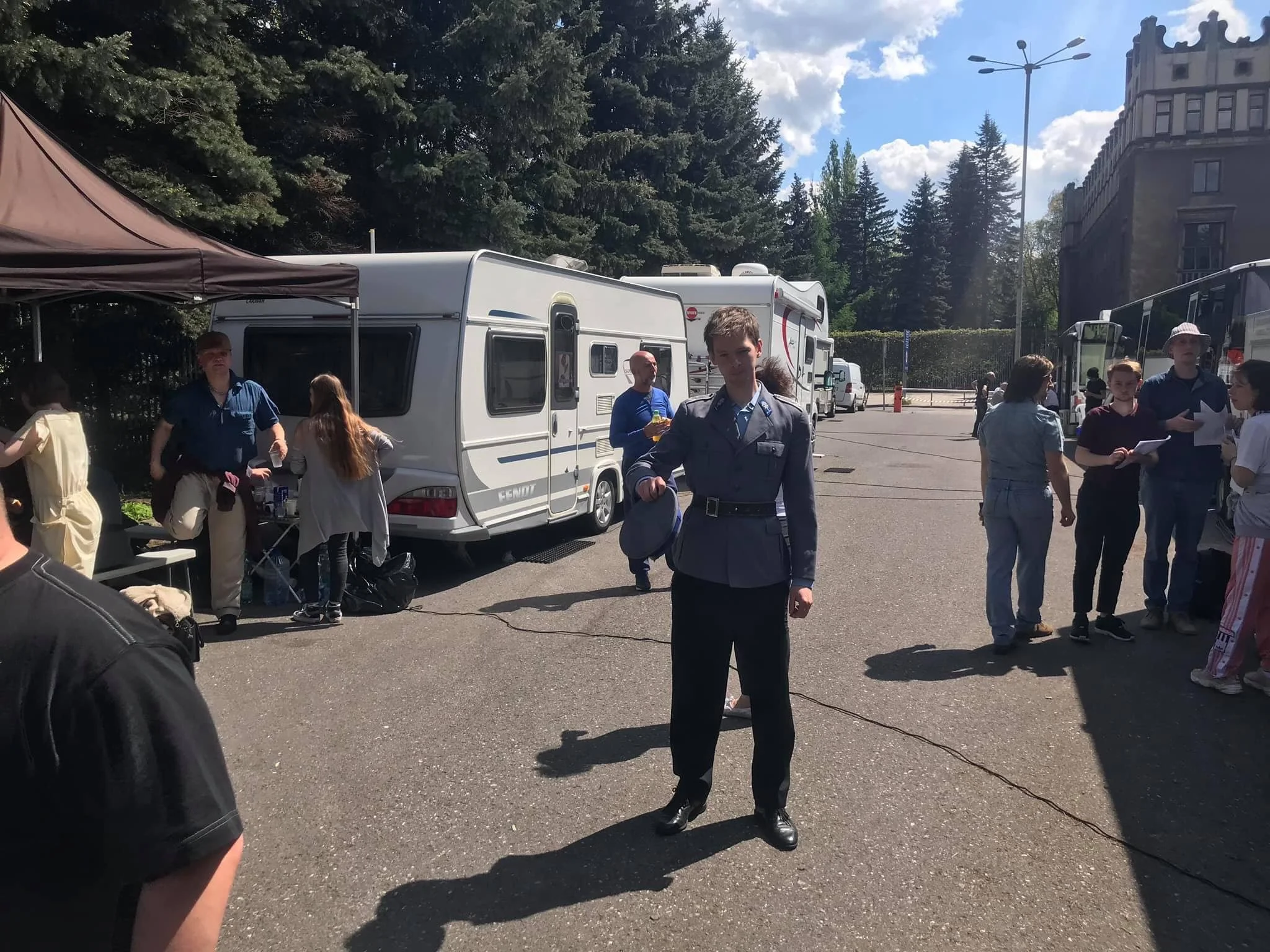 A woman in a police uniform standing on a parking lot with trees and a building in the background. Several people are gathered around, some looking at papers or talking, with caravans and buses visible.