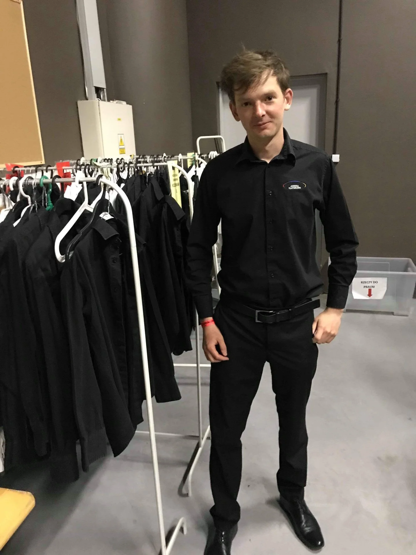 A young man wearing a black dress shirt and black pants standing next to a rack of black clothing in a backstage or storage room.