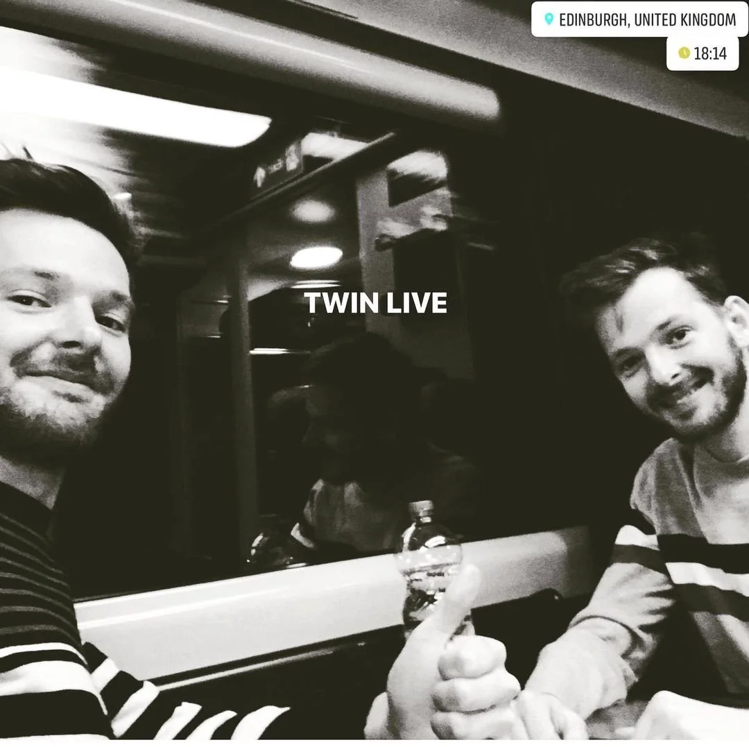 Two young men taking a selfie in a train, with one giving a thumbs up. Black and white photo with 'TWIN LIVE' written in the center. Location tag showing Edinburgh, United Kingdom, with the time 18:14.