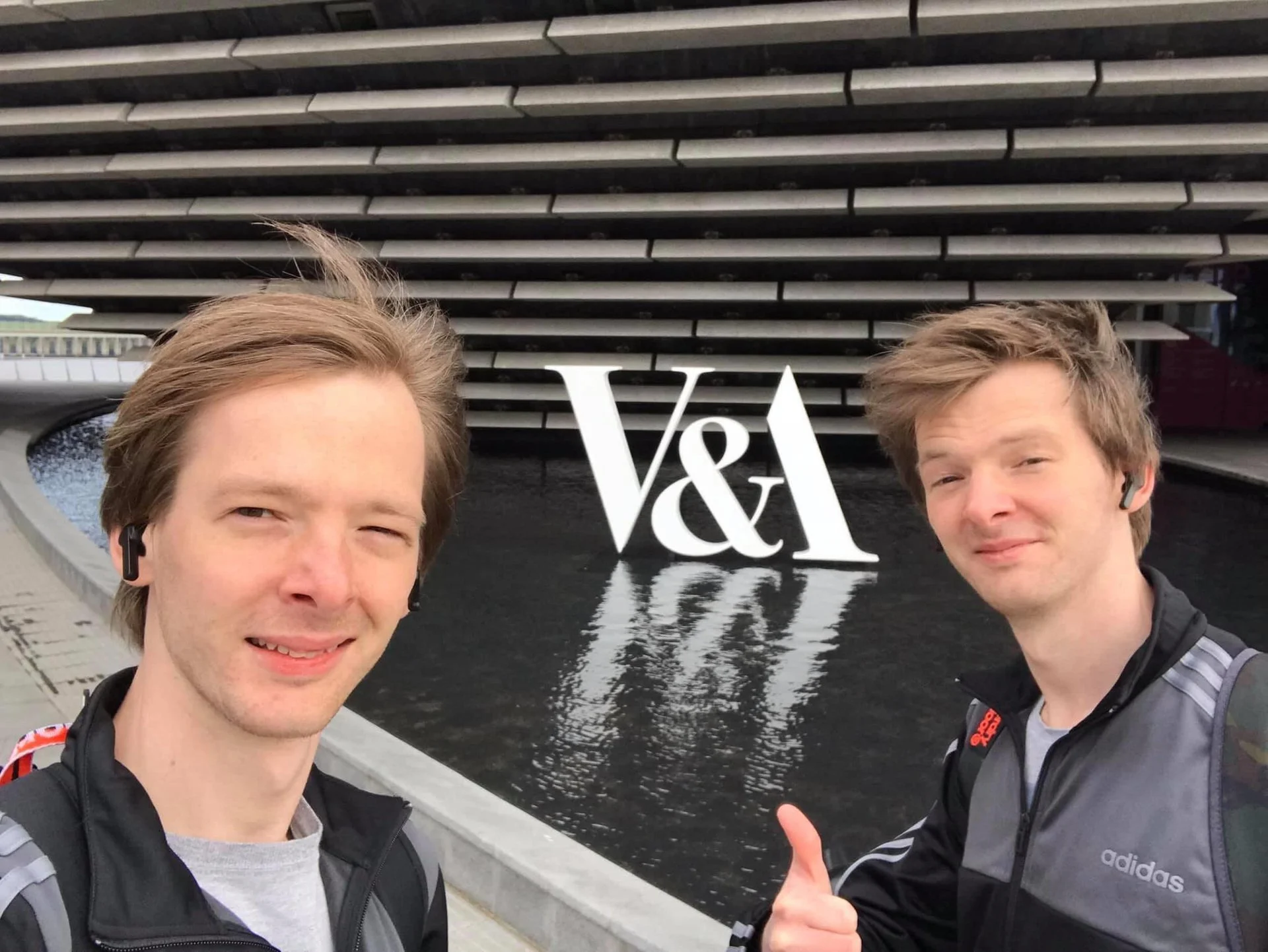 Two young men with light brown hair wearing black and gray Adidas athletic jackets and wireless earbuds, taking a selfie in front of a modern building with a water feature and a large white V&A sign, giving a thumbs-up.