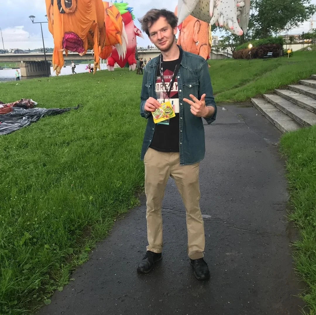 A young man standing on a paved pathway near a grassy area with a staircase to the right, holding a colorful candy package and making a peace sign gesture with his right hand. In the background, there are inflated colorful dragon and monster-shaped b