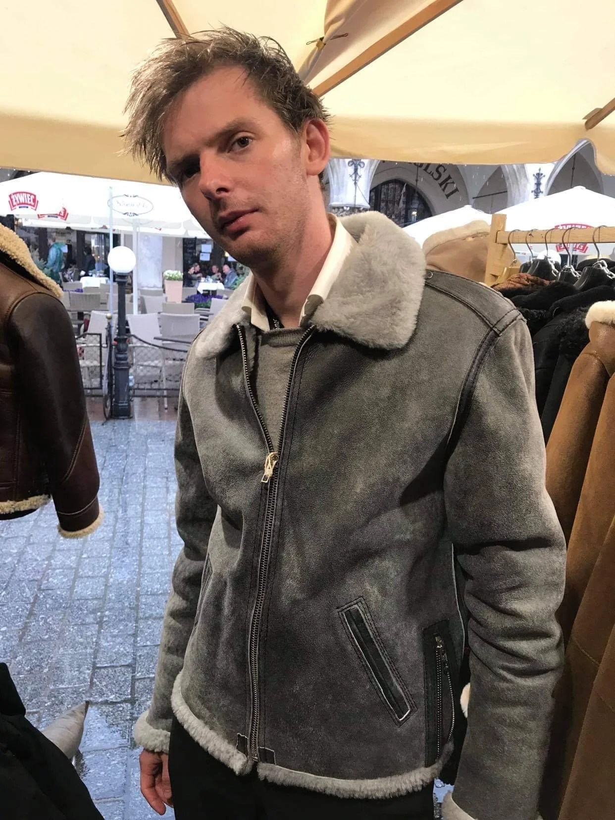 A young man with messy hair wearing a gray shearling-lined leather jacket standing outside a storefront at night, with outdoor seating and umbrellas visible in the background.