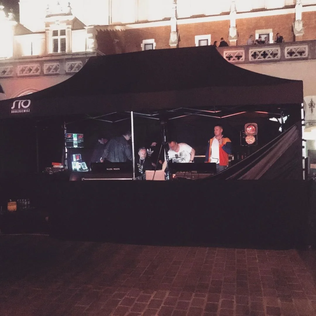 A live music performance at night under a black canopy tent with five musicians or performers on stage, surrounded by equipment, in an urban setting with illuminated historic-style buildings in the background.