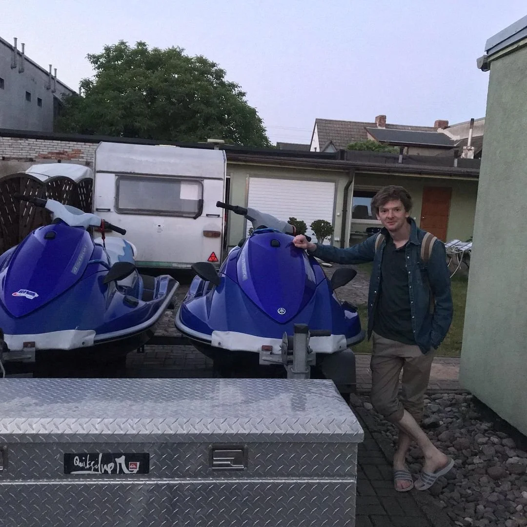 A young man standing outdoors next to two blue jet skis on a trailer, with a small caravan and residential buildings in the background.