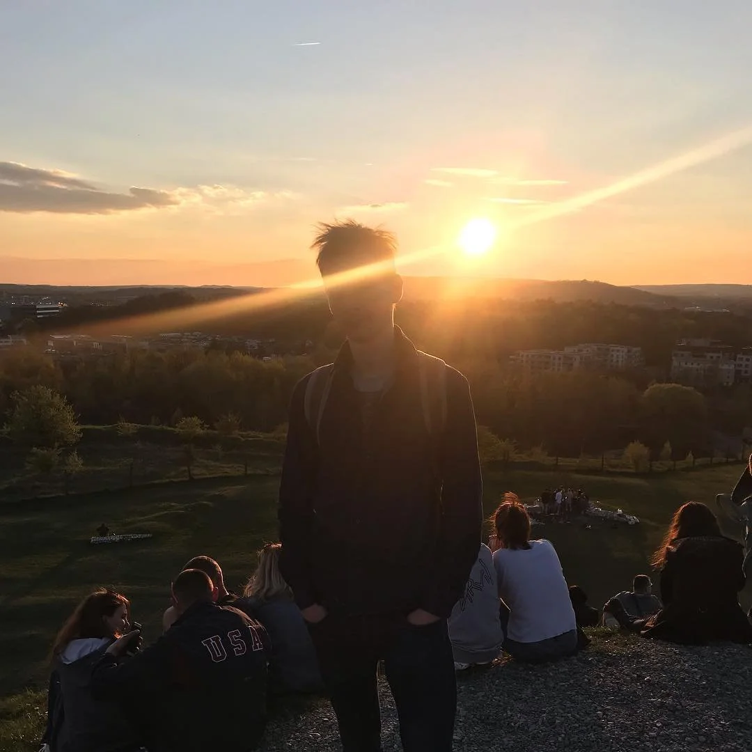 Person standing outdoors during sunset, silhouette with others sitting on a grassy hill in the background.