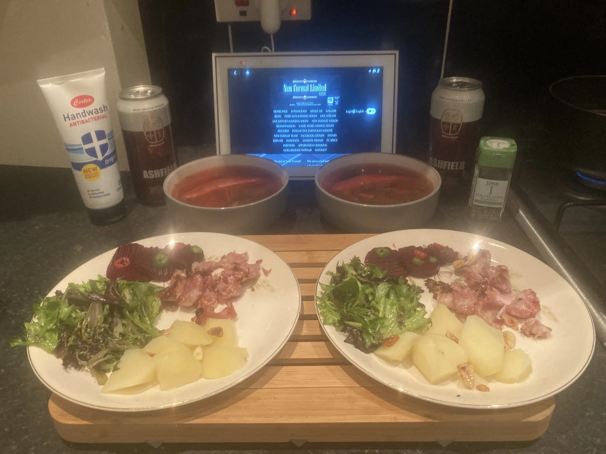 Two plates with salad, boiled potatoes, and slices of bacon, two bowls with soup, two cans of beer, a tube of hand wash, a small box of tea, and a tablet on a kitchen counter.