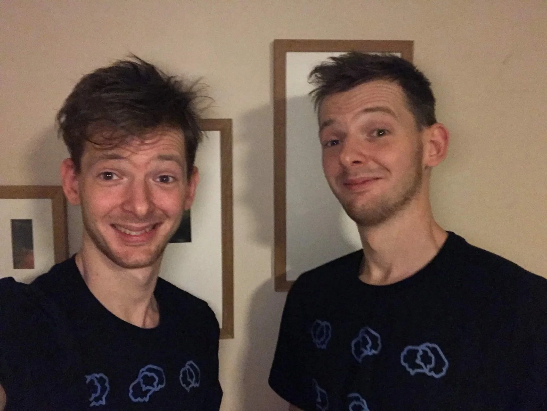 Two young men smiling and taking a selfie in a room with beige walls and framed pictures hanging on the wall behind them. They are both wearing black t-shirts with blue cloud patterns.