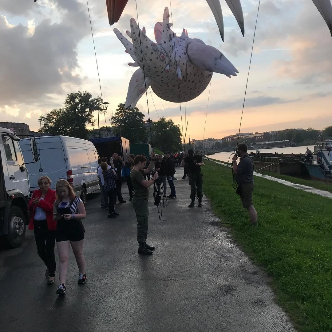 A large airship balloon shaped like a seagull floats in the sky near a river at sunset, with people on the ground taking photos and observing it.