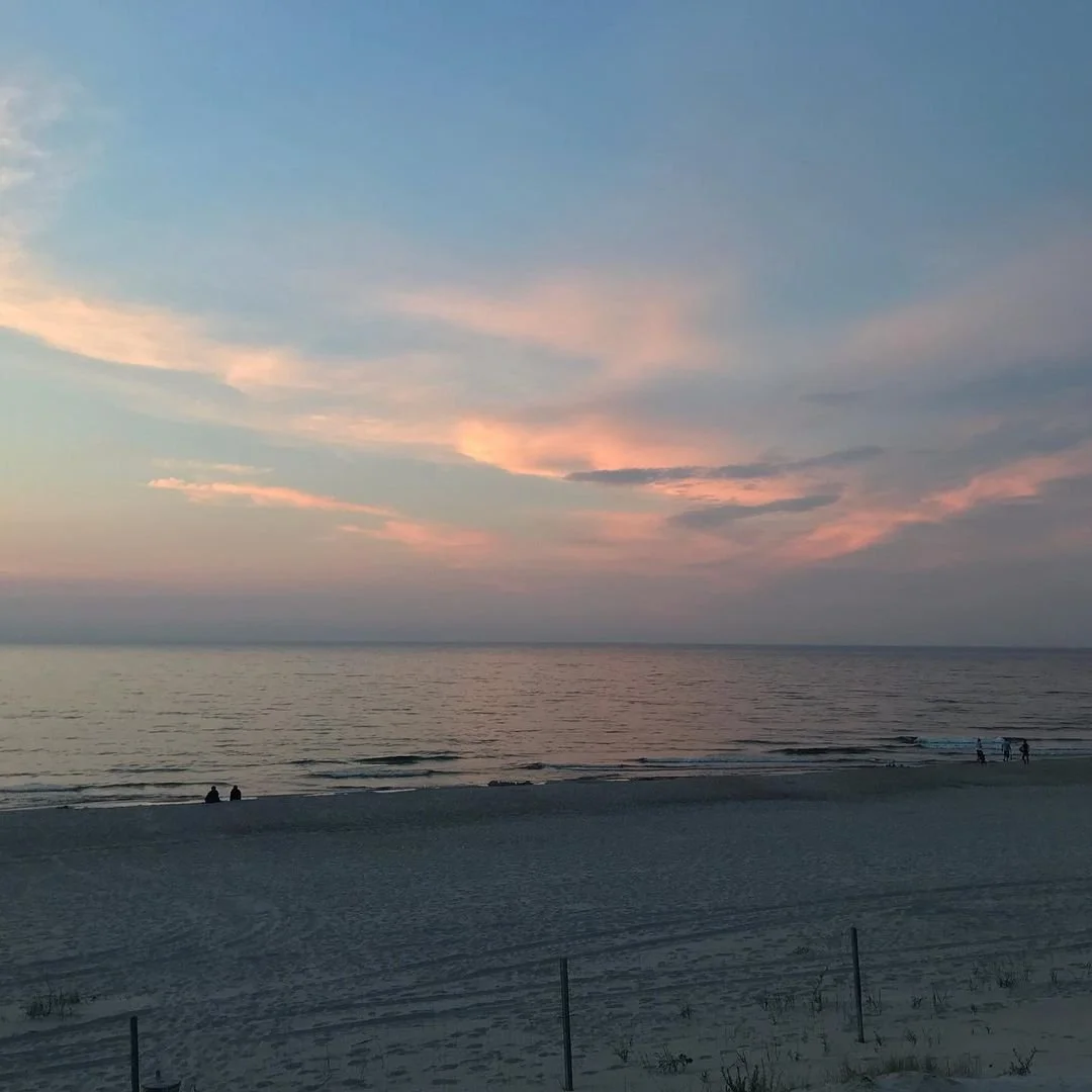 A peaceful beach scene at sunset with pink and blue clouds in the sky, calm ocean waves, and a few people sitting and walking along the sandy shore.