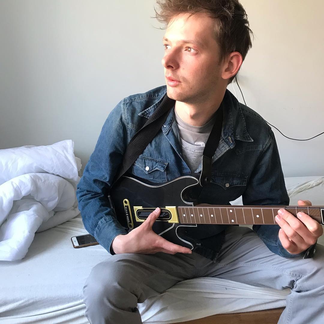 A young man sitting on a bed playing a small black electric guitar, with a smartphone lying beside him and a white pillow in the background.