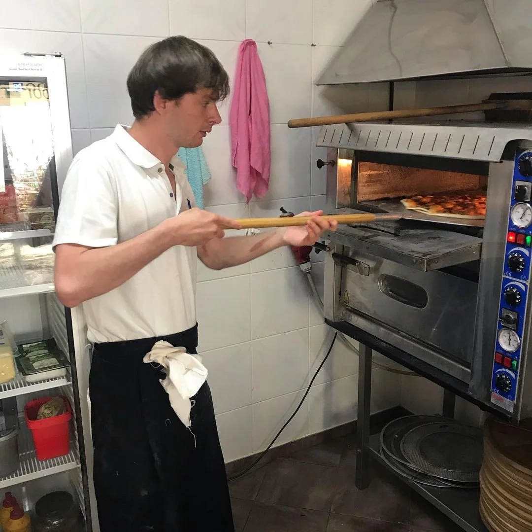 A man in a white polo shirt and black apron watches a pizza bake in an industrial oven, holding a long wooden pizza peel.