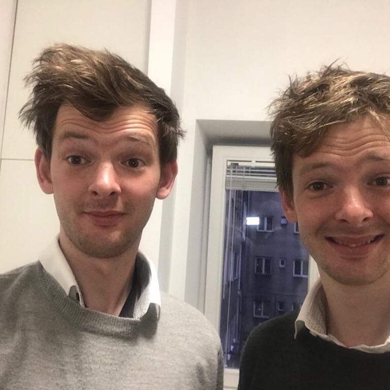 Two young men with messy hair taking a selfie indoors in front of a window at night.