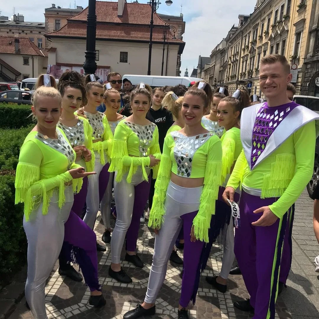 Group of performers wearing neon yellow and purple dance costumes with metallic silver accents, standing outdoors on a city street.