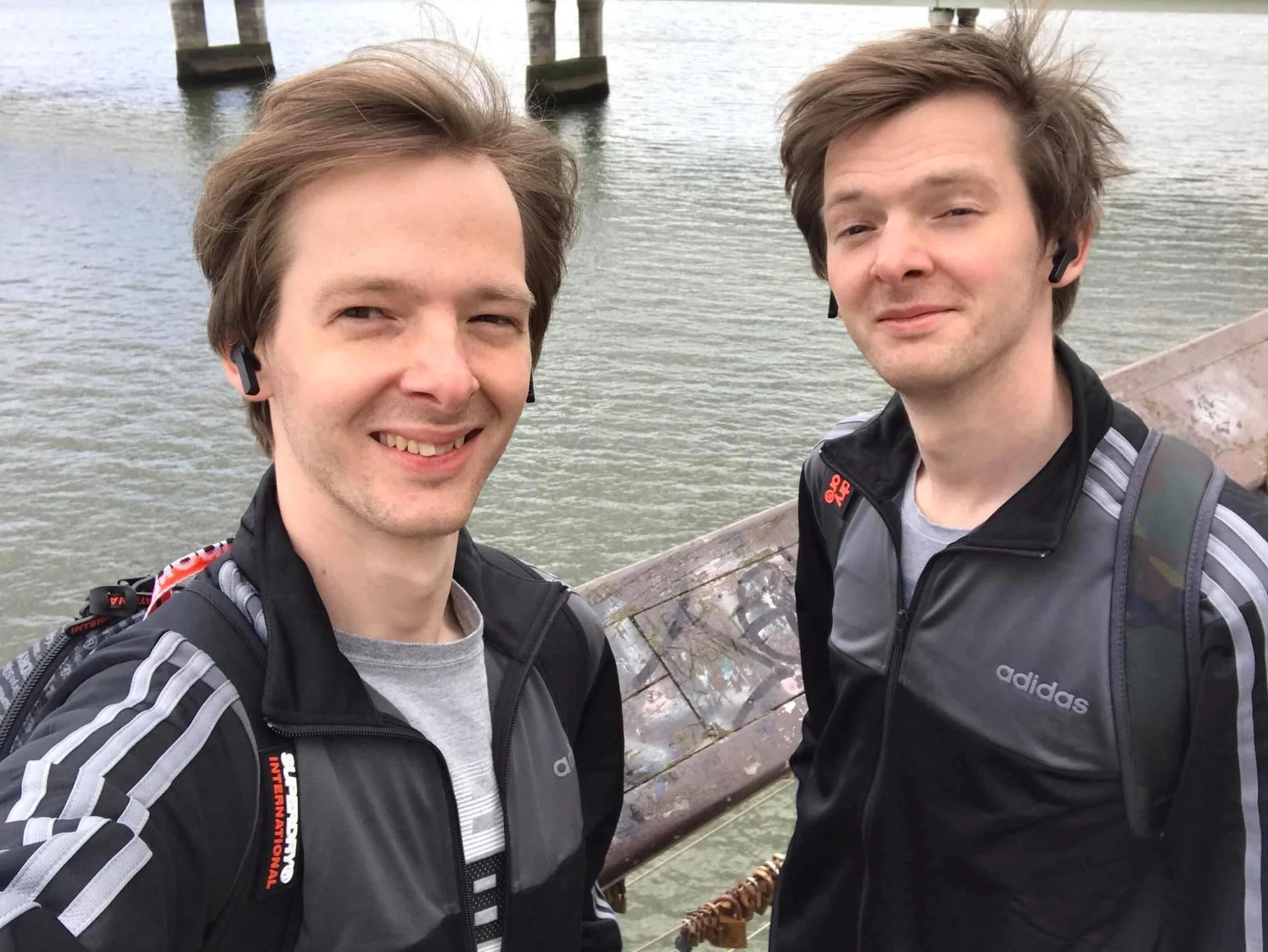 Two young men taking a selfie outdoors near water, both wearing black and gray Adidas jackets, backpacks, and wireless earbuds, smiling at the camera with a wooden dock or pier in the background.