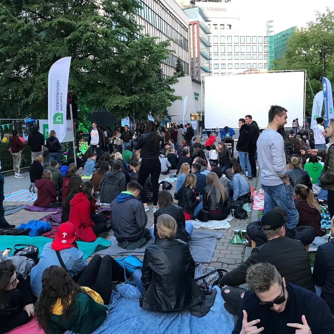 People sitting on blankets and standing in an outdoor urban area with a large screen, banners, and trees, at an event or outdoor movie screening.