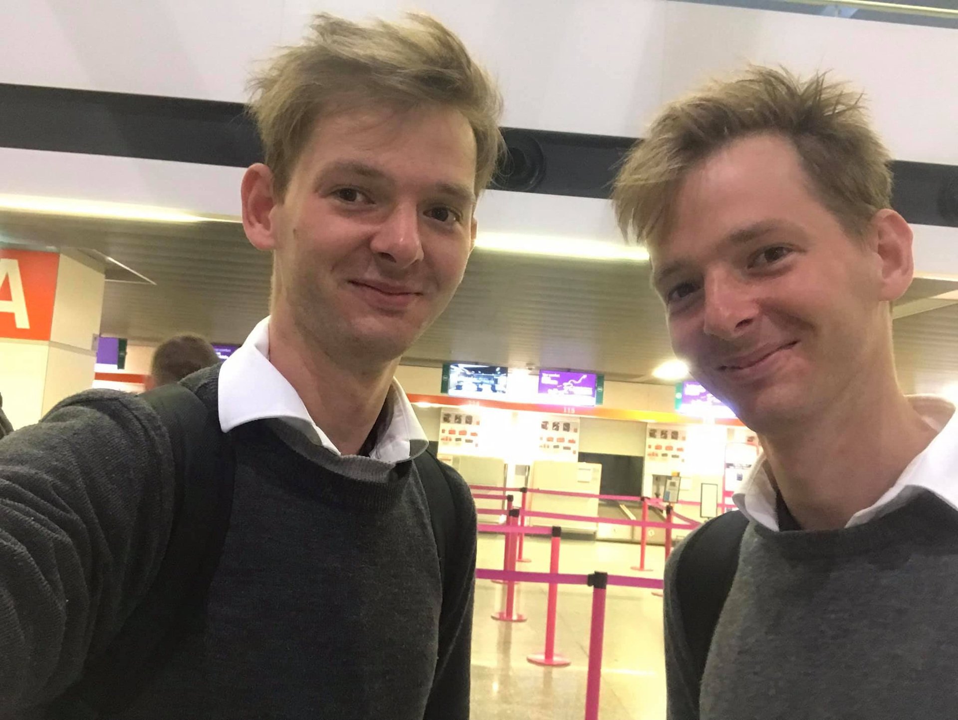 Two young men with light brown hair taking a selfie at an airport terminal, both wearing dark sweaters layered over white shirts, smiling at the camera.