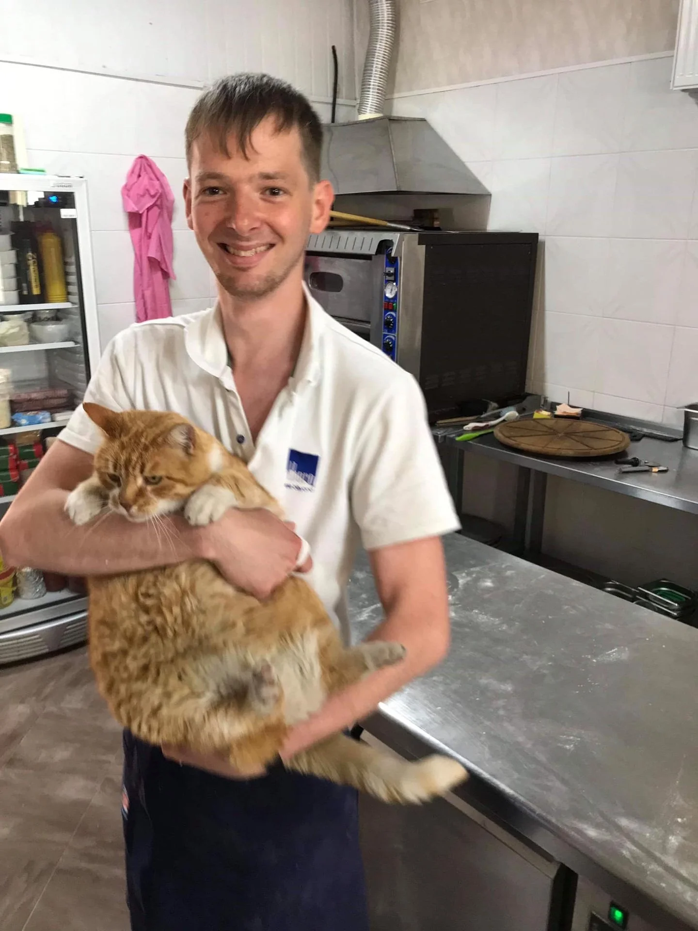 A man smiling and holding an orange tabby cat in a kitchen or bakery setting.