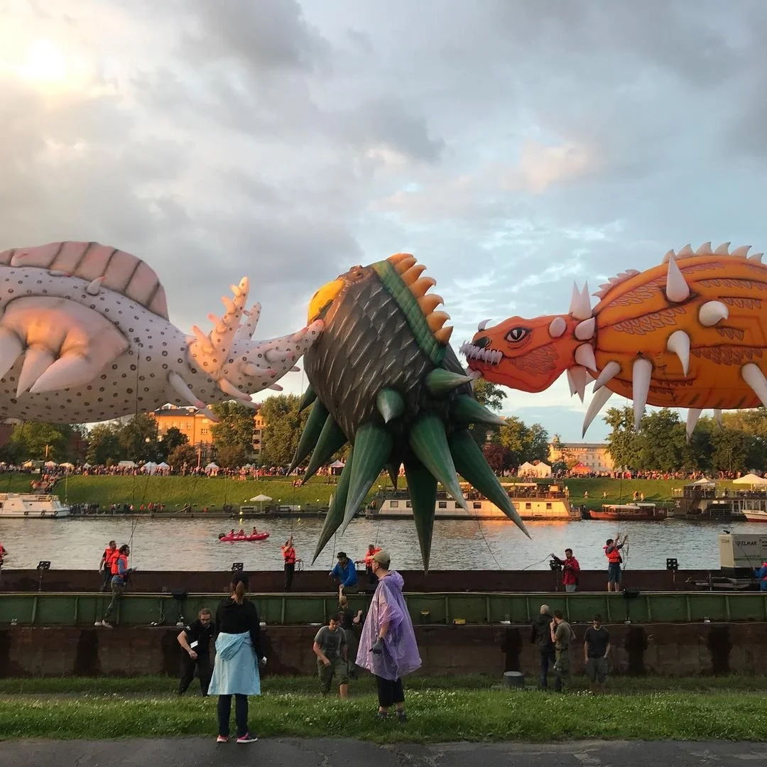 Colorful inflatable dragon and sea creature balloons floating over a river, with onlookers and a crowd in the background on a partly cloudy evening.