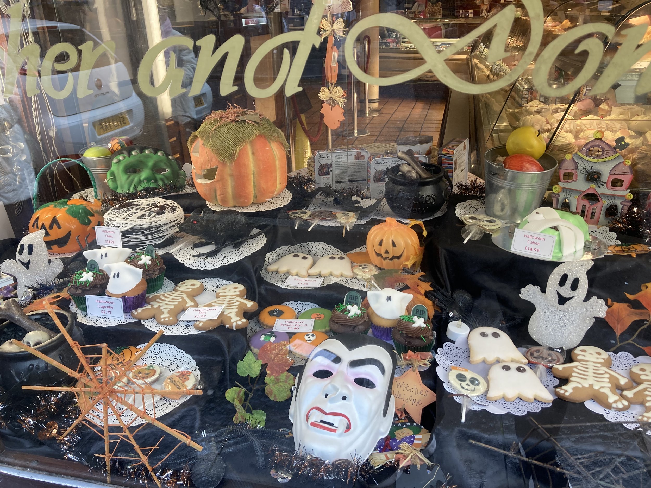 Display of Halloween-themed decorations and treats in a shop window, including ghost cookies, skeleton cookies, pumpkin decorations, witch's hat, ghost figures, a mask, and a spooky spider decoration.