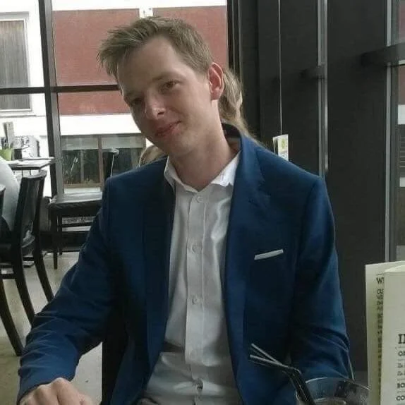 A young man with light brown hair, wearing a blue blazer and white shirt, sitting at a table in a restaurant with large windows and a view of a brick building outside.