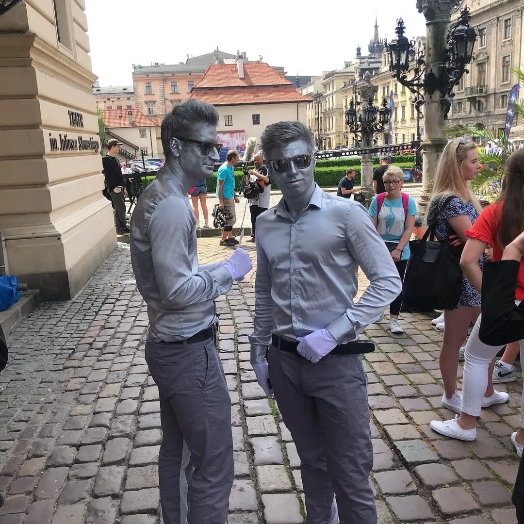 Two young men standing on a cobblestone street with painted faces, dressed in light gray shirts, sunglasses, and gloves, surrounded by a group of people in an urban setting with historic buildings.
