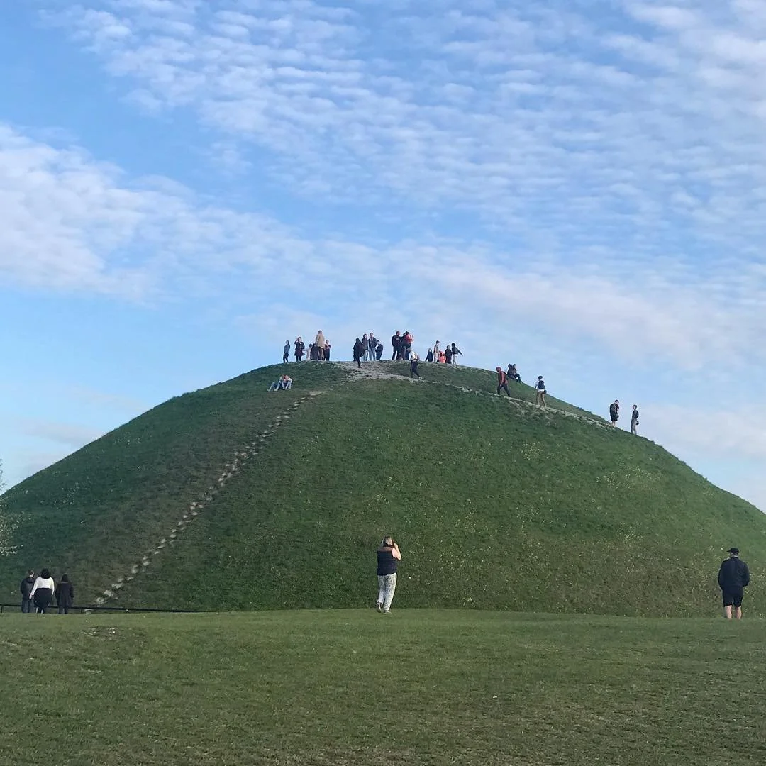 Green grassy hill with a worn narrow path leading to the top, where a group of people are standing and gathering, with some sitting on the hillside, under a partly cloudy blue sky.