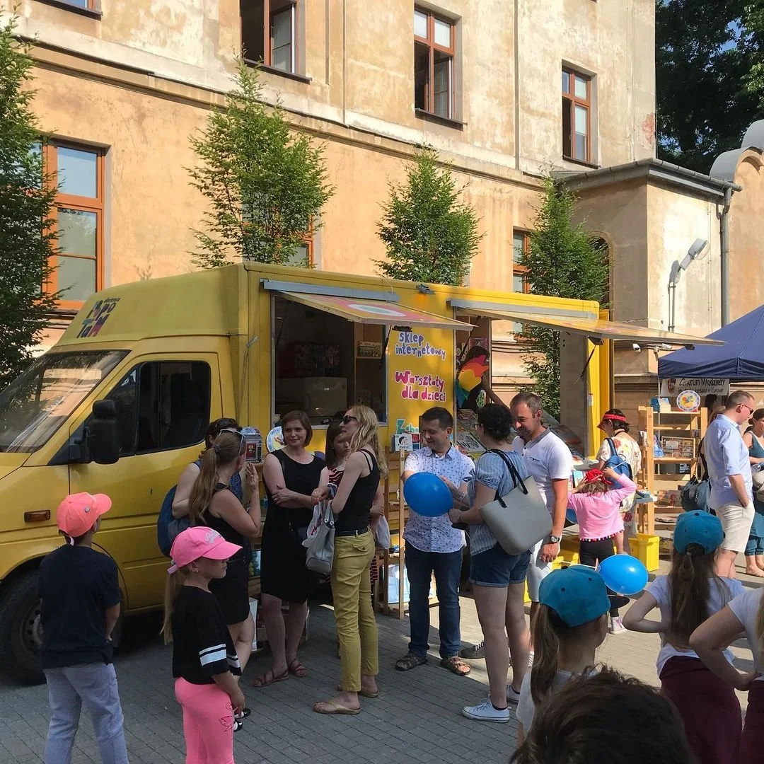 People gathered around a yellow mobile bookstore with open sides, selling books and children's activities, at an outdoor event.