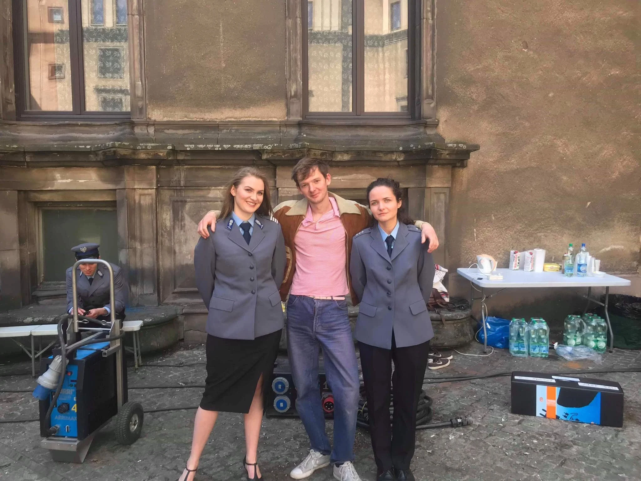 Three young adults, two women in police uniforms and a man in casual clothing, standing together with their arms around each other in front of an old building with large windows. There is a table with drinks and supplies on the right side and a woman