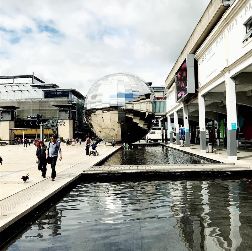 People walking near a reflective metallic spherical building with water feature in the foreground, in an urban area with modern architecture.
